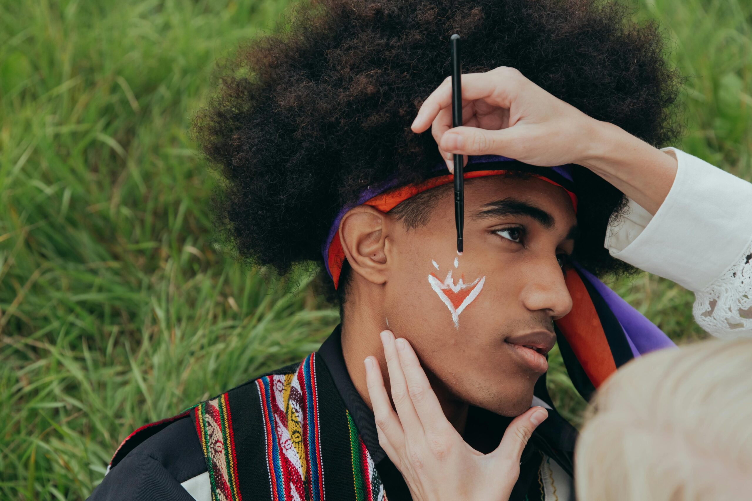 Young man with Afro hair getting his face painted outdoors, highlighting artistic expression and creativity.
