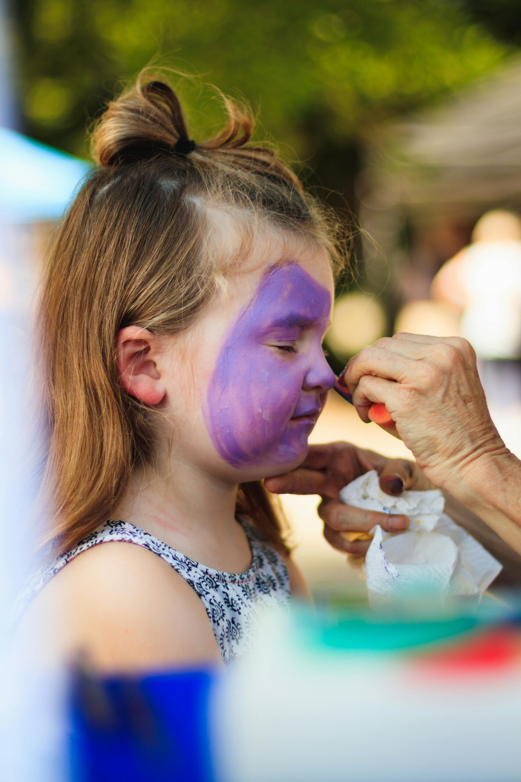 A young girl with blond hair is getting her face painted purple during an outdoor event on a sunny day.
